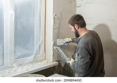 A Male Plasterer Installs A Perforated Paint Corner On The Corner Of The Window Slope And Plasters The Wall With A Spatula. 