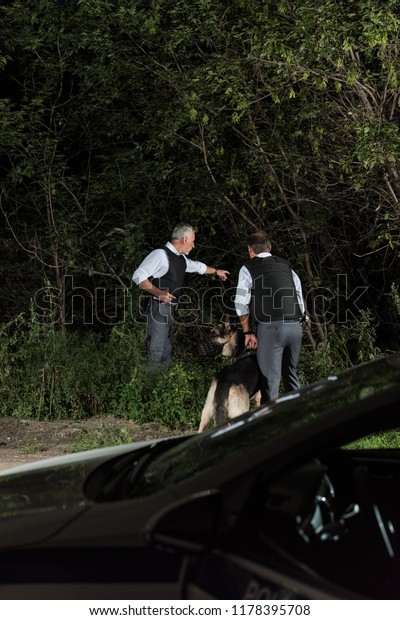 male police officer with gun\
pointing by finger to colleague with alsatian on leash outdoors\
