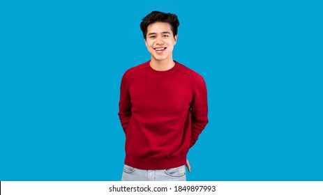 Male Portrait. Smiling asian guy standing and posing at studio, holding hands in pockets, looking at camera, wearing red sweater, posing isolated over blue studio background. Casual teenage model - Powered by Shutterstock