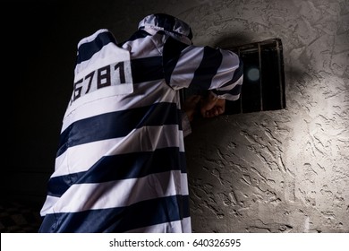 Male Prisoner Wearing Prison Uniform Tries To Escape Through The Window With Bars In A Small Dark Prison Cell