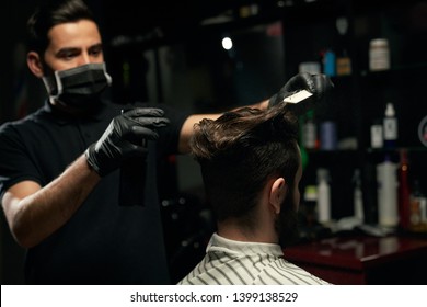 Male professional barber dressed in black shirt, wearing mask on mouth and gloves on hands is doing hair styling to unshaved man that covered with striped cape sitting in armchair at barbershop