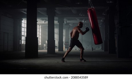 Male Professional Boxer Punching Sports Bag At Hadrcore Workout In Dark Gym. Angry Man Silhouette Working Hard Hit. African American Sportsman Powerful Body In Boxing Gloves Training At Sport Club