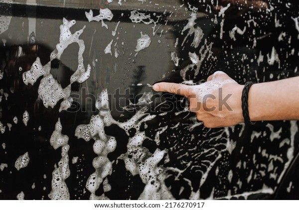 A male\
professional worker, a washerman, shows the result of his work with\
his finger on the background of a black dirty surface of a car door\
and white soap suds. Photography,\
concept.