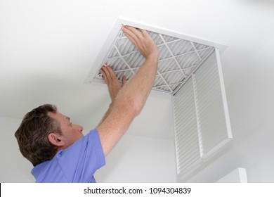 Male pushing a clean air filter into place in the ceiling with both hands. One fresh furnace air filter being secured in the intake grid of the white home ceiling.