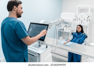 Male radiologist doing X-ray of female patient's hand on modern X-ray machine at hospital looking on computer screen. Patient wearing in protect lead apron. Medical examination of people - Powered by Shutterstock