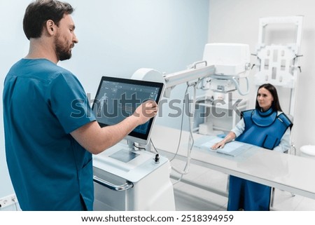 Male radiologist doing X-ray of female patient's hand on modern X-ray machine at hospital looking on computer screen. Patient wearing in protect lead apron. Medical examination of people