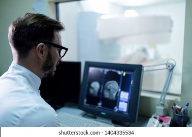 Male Radiologist Reviewing At Brain MRI Scan Results On The Computer In Control Room At Clinic. 