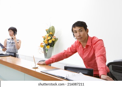 Male receptionist leaning on desk, colleague in background, portrait