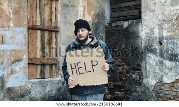Male refugee in hat cap is in foreign country\
because of war in his own country. Illegal caucasian immigrant\
stands outside an abandoned building looking for job. Consequences\
of drunkenness, alcoholi