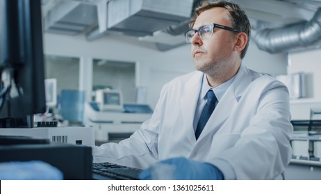 Male Research Scientist Sits at His Workplace in Laboratory, Uses Personal Computer. I the Background Genetics, Pharmaceutical Research Centre with Innovative Equipment.