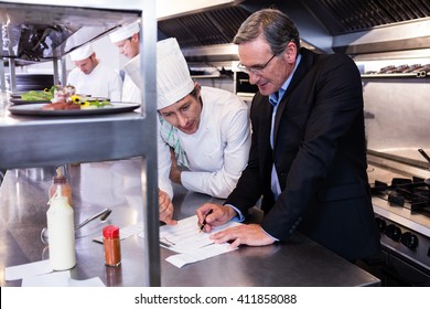 Male Restaurant Manager Writing On Clipboard While Interacting To Head Chef In Commercial Kitchen