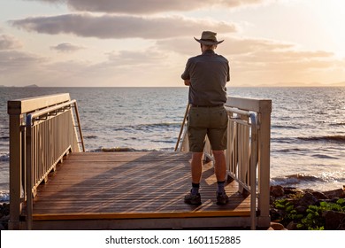 A Male Retiree In His Seventies Standing On A Timber Deck With Stairs Leading Down To The Beach. He's Looking Out To See, Watching The Waves And Admiring The Early Morning Light