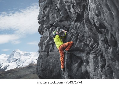 Male Rock Climber. Rock Climber Climbs On A Black Rocky Wall On The Ocean Bank In Iceland, Kirkjufjara Beach. Man Makes Hard Move Without Rope.