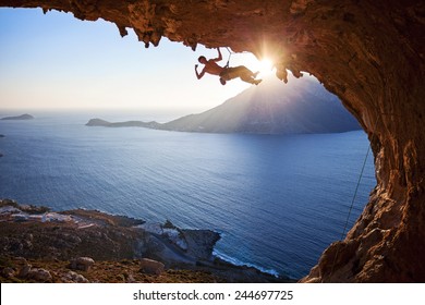 Male Rock Climber Flexing His Bicep While Hanging On One Arm At Sunset. Climbing Along Roof In Cave, Kalymnos, Greece.