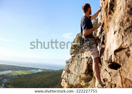 A male rockclimber climbing up a steep mountain attached to a rope and harness looking at the view