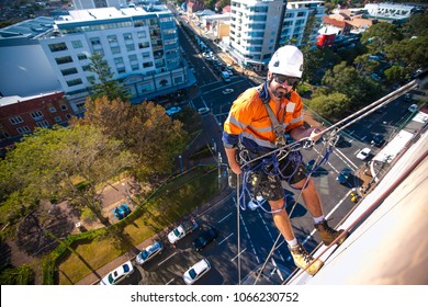 Male Rope Access Industry Worker Wearing Full Body Safety Harness, White Hard Hat, Using Static Twin Ropes Working Abseiling From Construction High Rise Building Site In Sydney City, Australia  