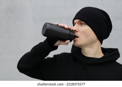 Male Runner Drinking Water From A Black Plastic Bottle After A Cardio Workout. Sportsman In Black Hoodie And Cap Hydrating During Outdoor Training.