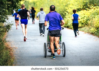 Male Runner Running Marathon In Autumn City Park With A Pram Baby