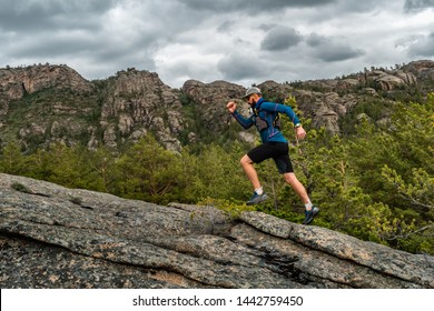 Male Runner Running On A Mountain Trail. Athlete Runs In The Mountains Among The Rocks. Man In Blue Jersey And Black Shorts Training Outdoors