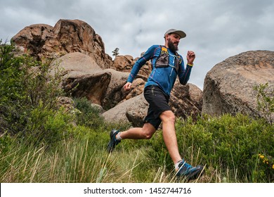 Male Runner Running On A Mountain Trail. Athlete Runs In The Mountains Among The Rocks. Man In Blue Jersey And Black Shorts Training Outdoors