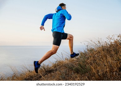 male runner running uphill in blue jacket and black tights, background of sky and sea, trail dry grass - Powered by Shutterstock