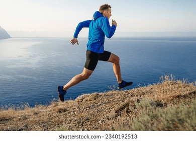 male runner running uphill trail on edge in blue jacket and black tights, background of sky and sea