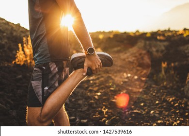 Male Runner Stretching Leg And Feet And Preparing For Running Outdoors. Smartwatches Or Fitness Tracker On Hand. Beautiful Sun Light On Background. Active And Healthy Lifestyle Concept.