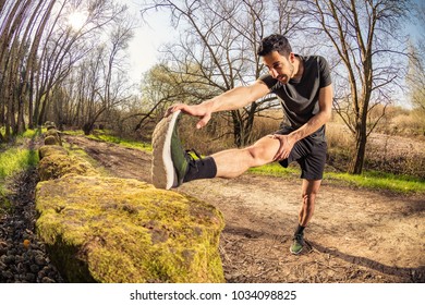 Male Runner Stretching Outdoors On Nature In Misty Forest Full Of Pleasure Warm Light. Standing Hamstring Stretch On Old Stone Wall.