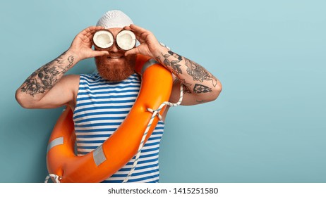 Male safeguard supervises situation on sea, holds two coconuts on eyes instead of binoculars, wears white swimhat, sailor vest, uses safety equimpent like ring buoy, poses over blue wall, blank space