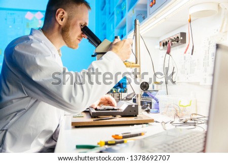 Male scientist wearing robe uses microscope for examining and soldering components on electronic cirquit board.
