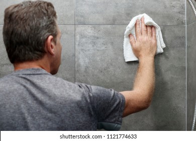 Male Scrubbing A Home Bathroom Gray Tile Shower Wall With His Hand Holding A Wet White Cotton Rag. White Cotton Cloth Being Used By An Older Caucasian Man To Clean A Bathroom Shower Gray Tile Wall.