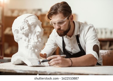Male sculptor repairing gypsum sculpture of woman's head at the working place in the creative artistic studio.
