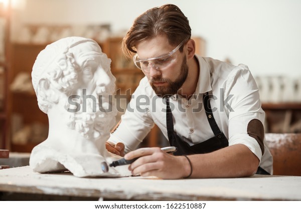 Male sculptor\
repairing gypsum sculpture of woman\'s head at the working place in\
the creative artistic\
studio.
