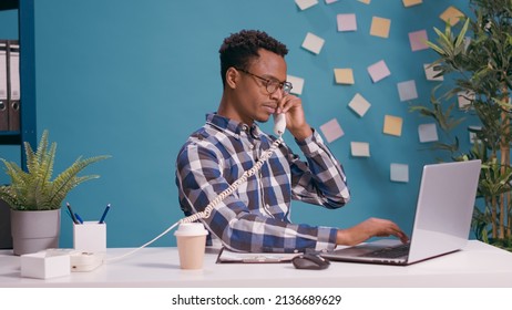 Male secretary answering landline phone at desk with laptop, using office vintage telephone to speak to people. Call center operator talking on phone call to give telecommunications support.