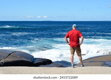 A male senior wearing a red shirt, blue hat and tan shorts standing on the edge of a rocky shore with an angry ocean on a sunny day. The waves are crashing against the coastline's trail and splashing. - Powered by Shutterstock