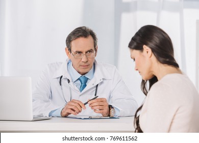 Male Serious Doctor And Female Patient Sitting At Talking At Clinic