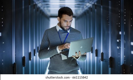 Male Server Engineer Works on a Laptop in Large Data Center.