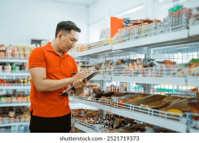 male shop assistant stands writing and checking a list on a clipboard while working at a minimarket - Powered by Shutterstock