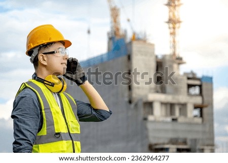 Male site engineer or foreman using mobile phone talking with his coworker team. Asian worker man with green reflective vest, safety helmet, goggles and earmuffs working at construction site
