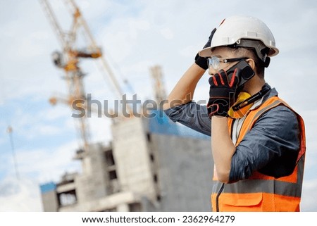 Male site engineer or foreman using mobile phone talking with his coworker team. Asian worker man with orange reflective vest, safety helmet, goggles and earmuffs working at construction site