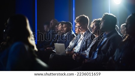 Male Sitting in a Dark Crowded Auditorium at a Tech Conference. Professional Using Laptop Computer. Specialist Watching Innovative Technology Presentation About New Hardware and High Tech Solutions.