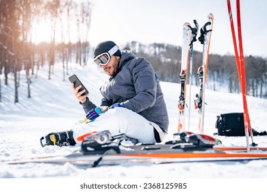Male skier sitting on snow taking a break while using phone. Man wearing grey and white ski suit enjoying winter holidays outdoors.