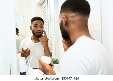 Male Skin Care. Handsome African Man Applying Moisturizing Cream In Bathroom