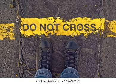 Male Sneakers On The Asphalt Road With Yellow Line And Title Do Not Cross. Border Line Concept, Danger Or Warning Sign, Top View