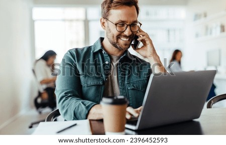 Male software designer speaking to a client on the phone while using a laptop. Happy young business man working in a co-working office.