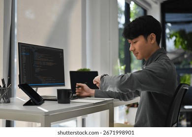 A Male software developer checking his smartwatch while working at a modern desk with code displayed on a monitor.