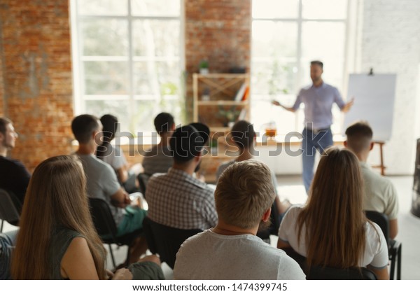 Male speaker giving presentation in hall at\
university workshop. Audience or conference hall. Rear view of\
unrecognized participants in audience. Scientific conference event,\
training. Education