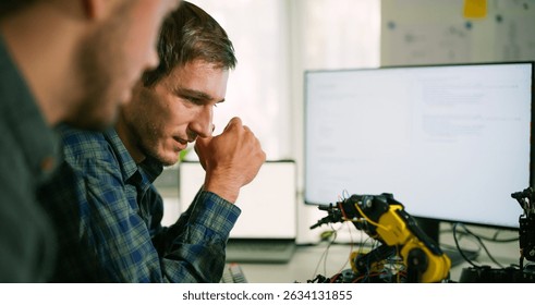 Male specialist engineers analyze code and robotic arm behavior during testing phase of an automation project in a high-tech workspace.