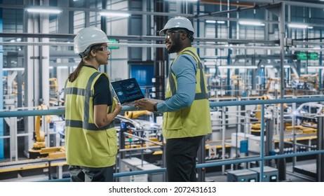 Male Specialist And Female Car Factory Engineer In High Visibility Vests Using Laptop Computer. Automotive Industrial Manufacturing Facility Working On Vehicle Production. Diversity On Assembly Plant.