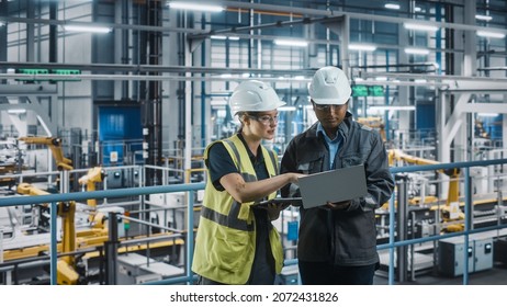 Male Specialist And Female Car Factory Engineer Talking And Working On Laptop And Tablet Computers. Automotive Industry 4.0 Facility Working On Vehicle Production. Diversity On Assembly Plant.